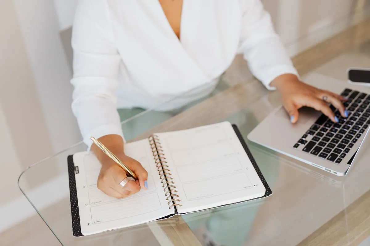Host writing notes in a planner while typing on a laptop, comparing AI draft replies with her own answers to guests